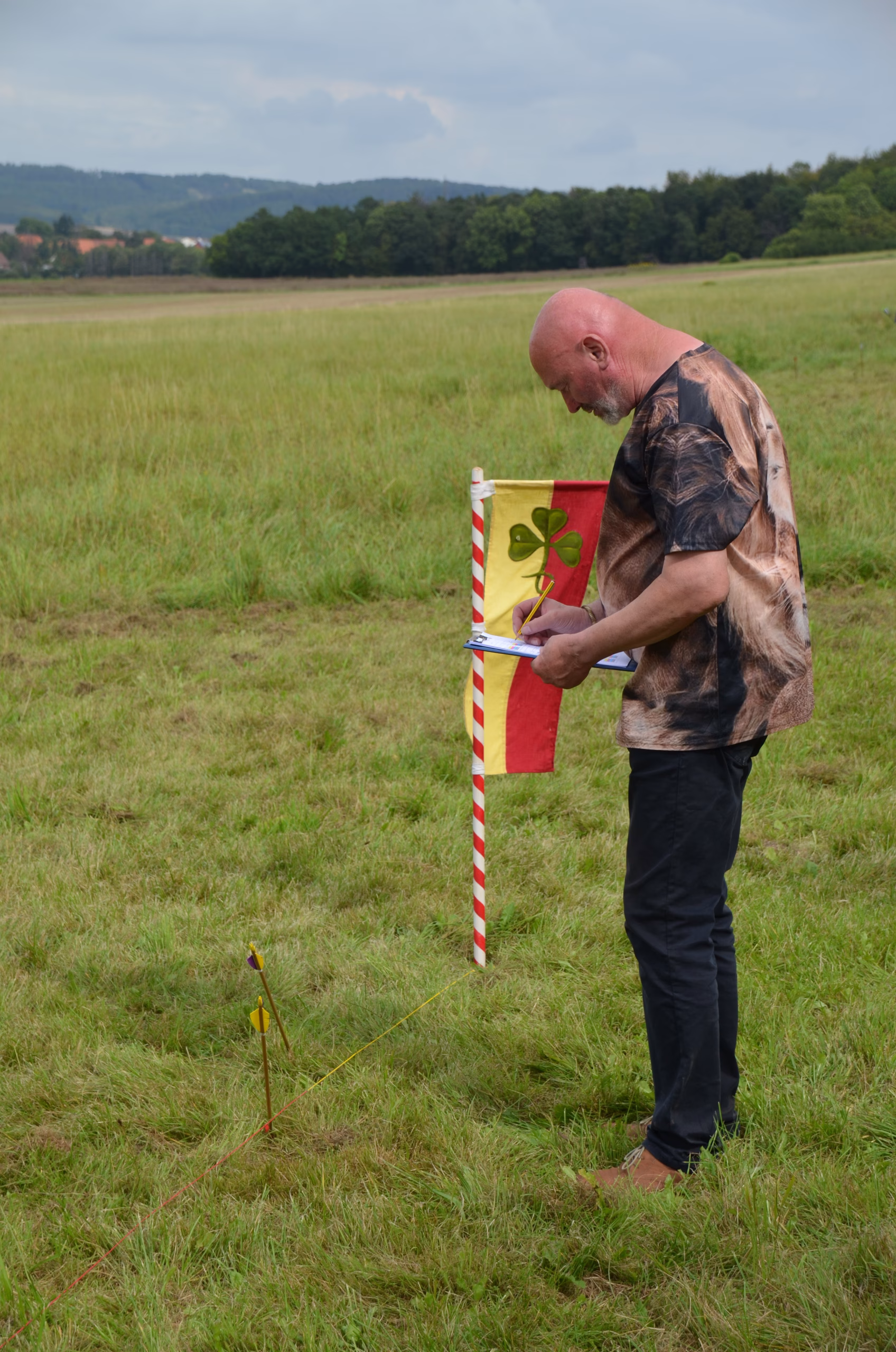 Bogenschütze notiert Wertung beim Clout-Bogenschießen in Siebenberge, nahe der Ziel-Flagge und eingeschlagenen Pfeilen.