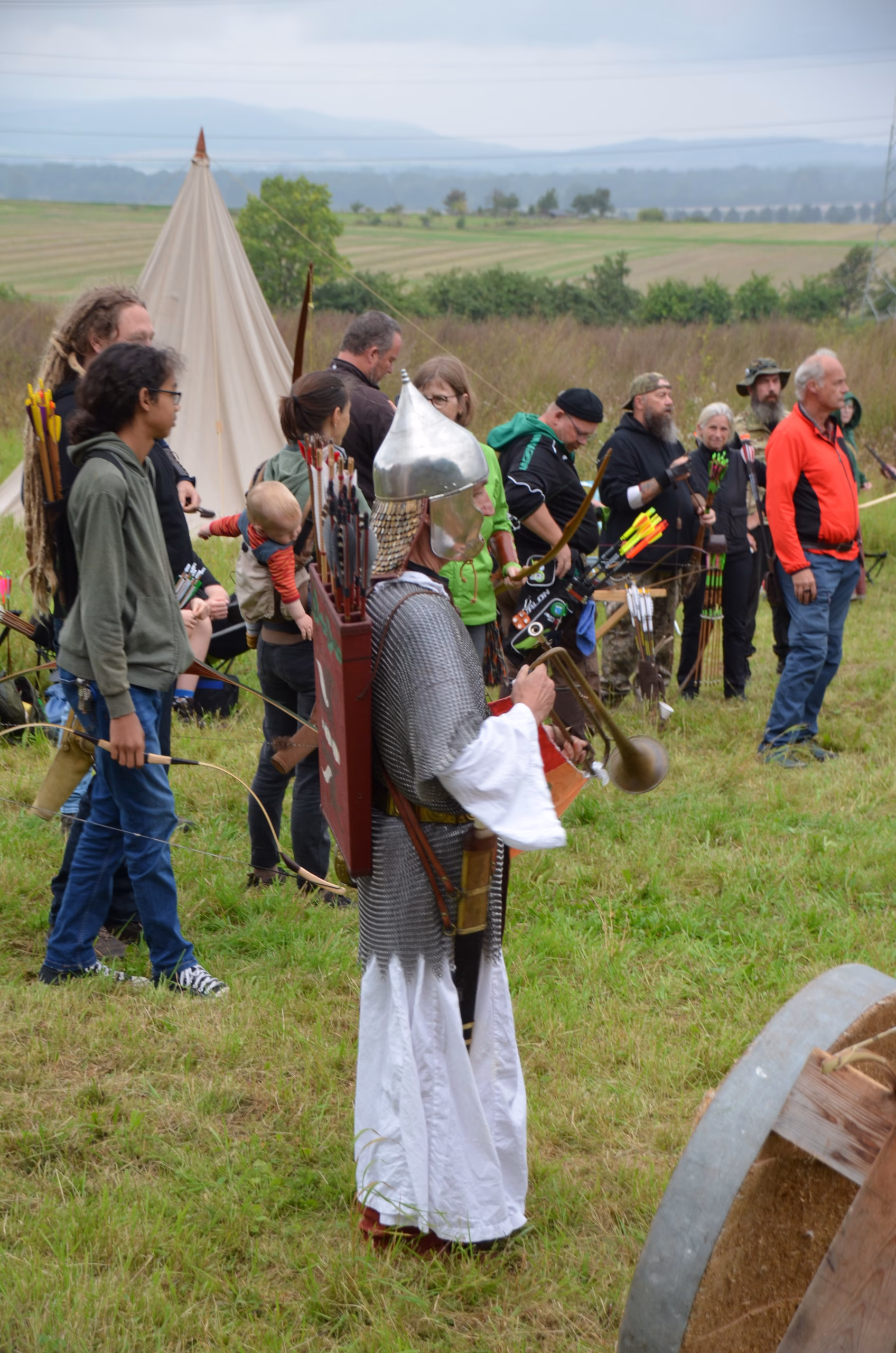 Bogenschützen beim Clout-Wettbewerb in Siebenberge, 2021. Ein Teilnehmer in historischer Rüstung mit Helm und Kettenhemd steht im Vordergrund, weitere Bogenschützen und Zuschauer sind im Hintergrund zu sehen. Veranstalter: Pfeilflugkunst.