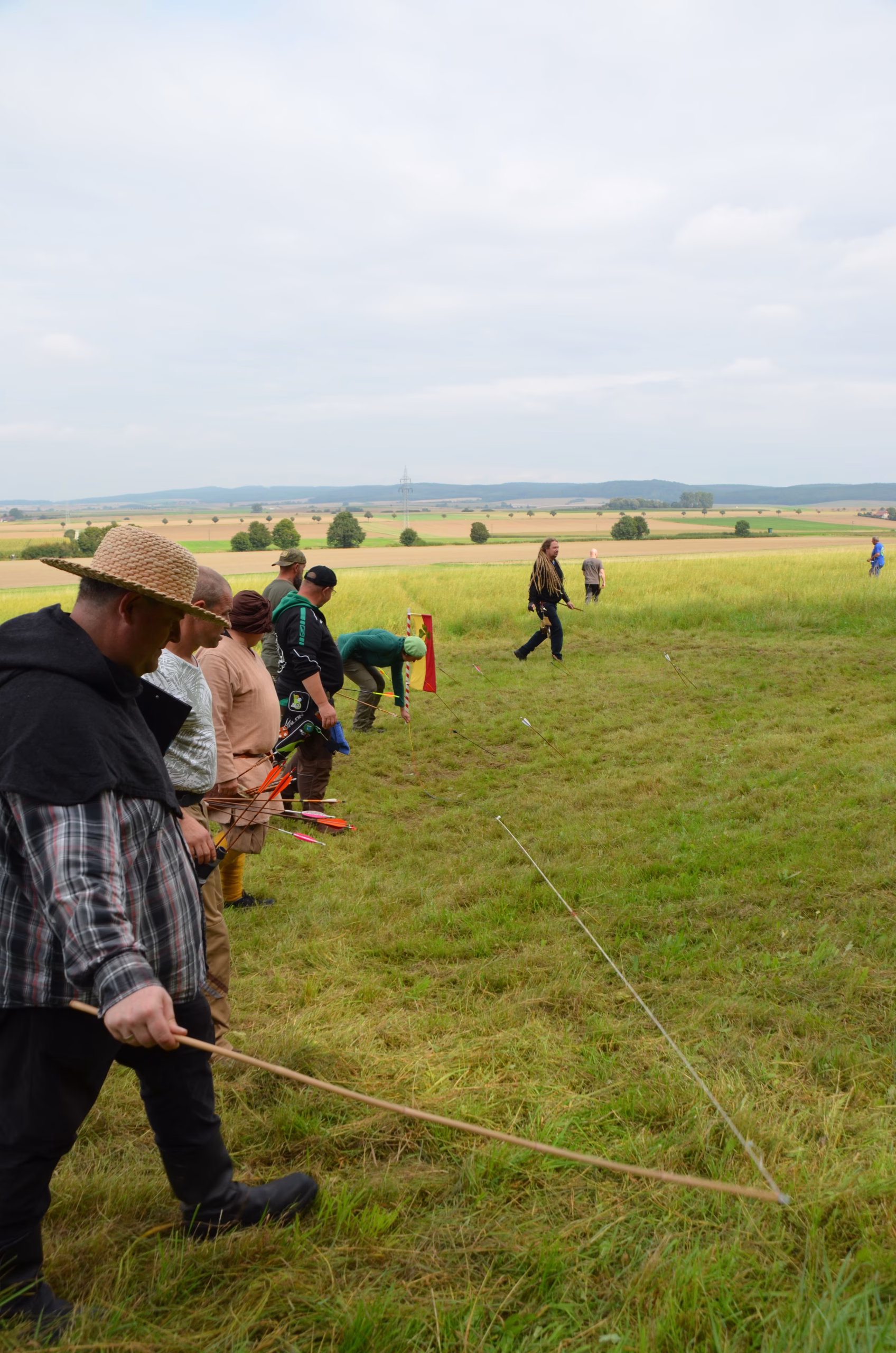 Gruppe von Bogenschützen beim Siebenberge Clout Bogenwettbewerb 2021 von Pfeilflugkunst, mit Pfeilen im Gras und Clout-Flagge im Hintergrund.
