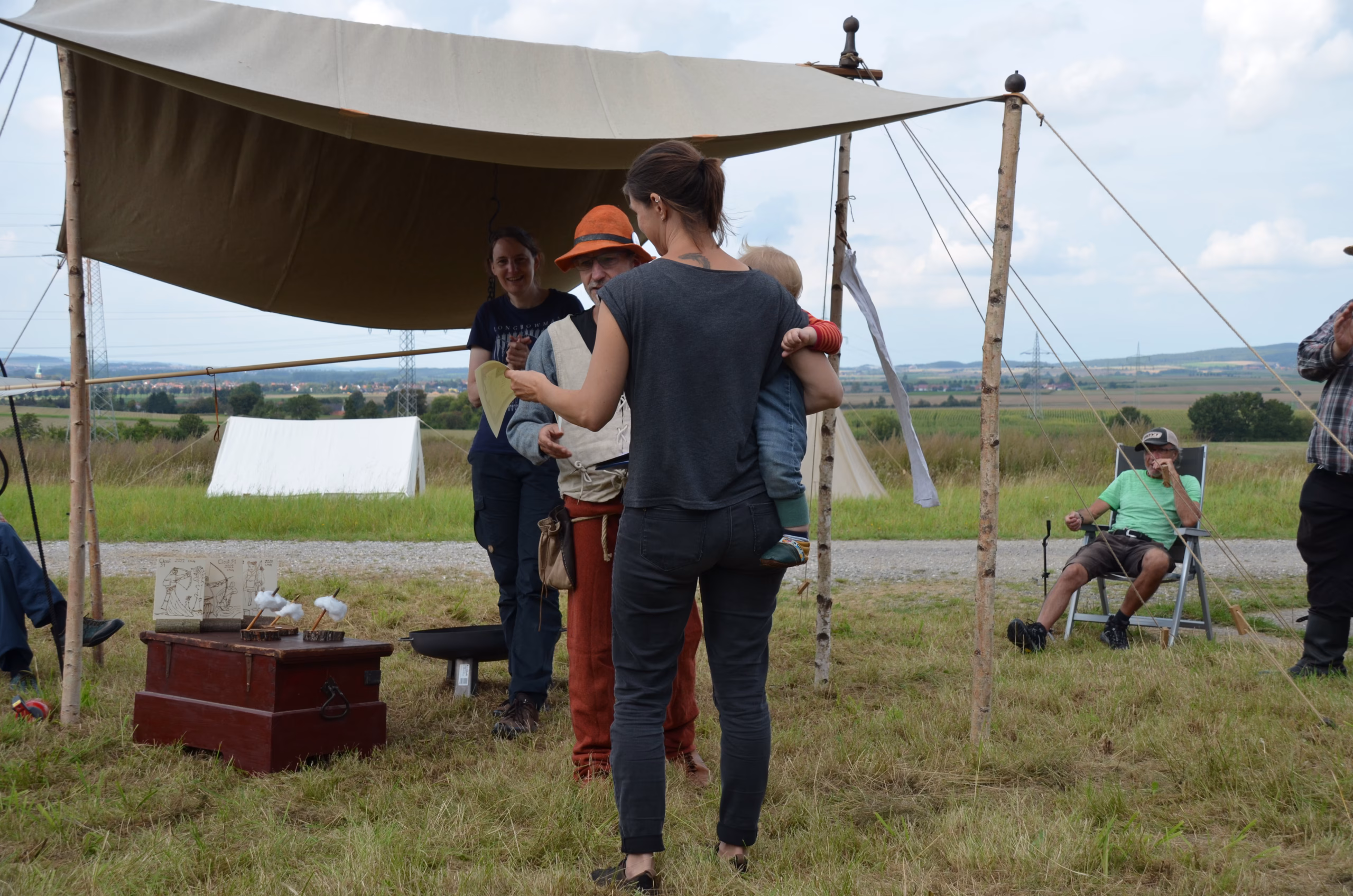 Teilnehmer des Siebenberge Clout Bogenwettbewerbs 2021 von Pfeilflugkunst unter einem Zelt, mit einer Frau, die ein Kind hält, und einem Mann in historischer Kleidung bei einer Besprechung oder Siegerehrung in den Siebenbergen.