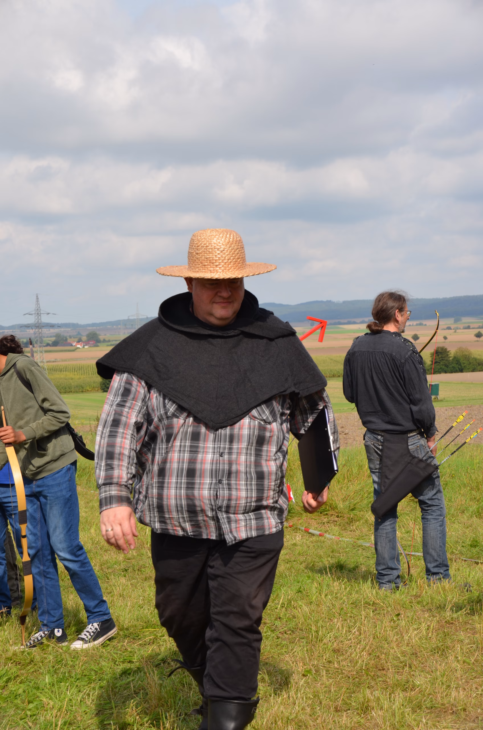 Ein Mann mit Strohhut und Umhang geht über ein Feld beim Clout Bogenwettbewerb in Siebenberge, veranstaltet von Pfeilflugkunst.