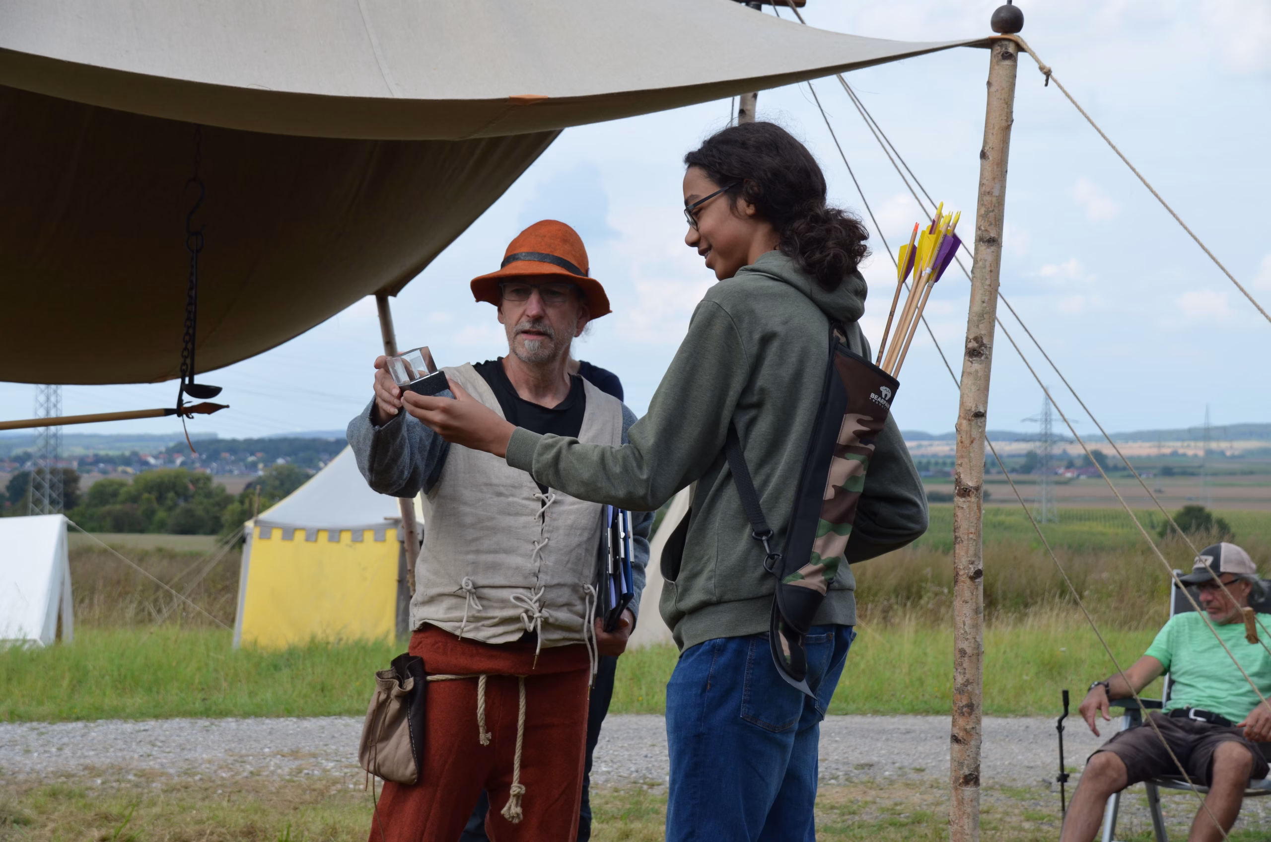 Zwei Bogenschützen, ein älterer in traditioneller Kleidung und ein junger mit Köcher, unterhalten sich unter einem Zelt beim Siebenberge Clout Bogenwettbewerb 2021.