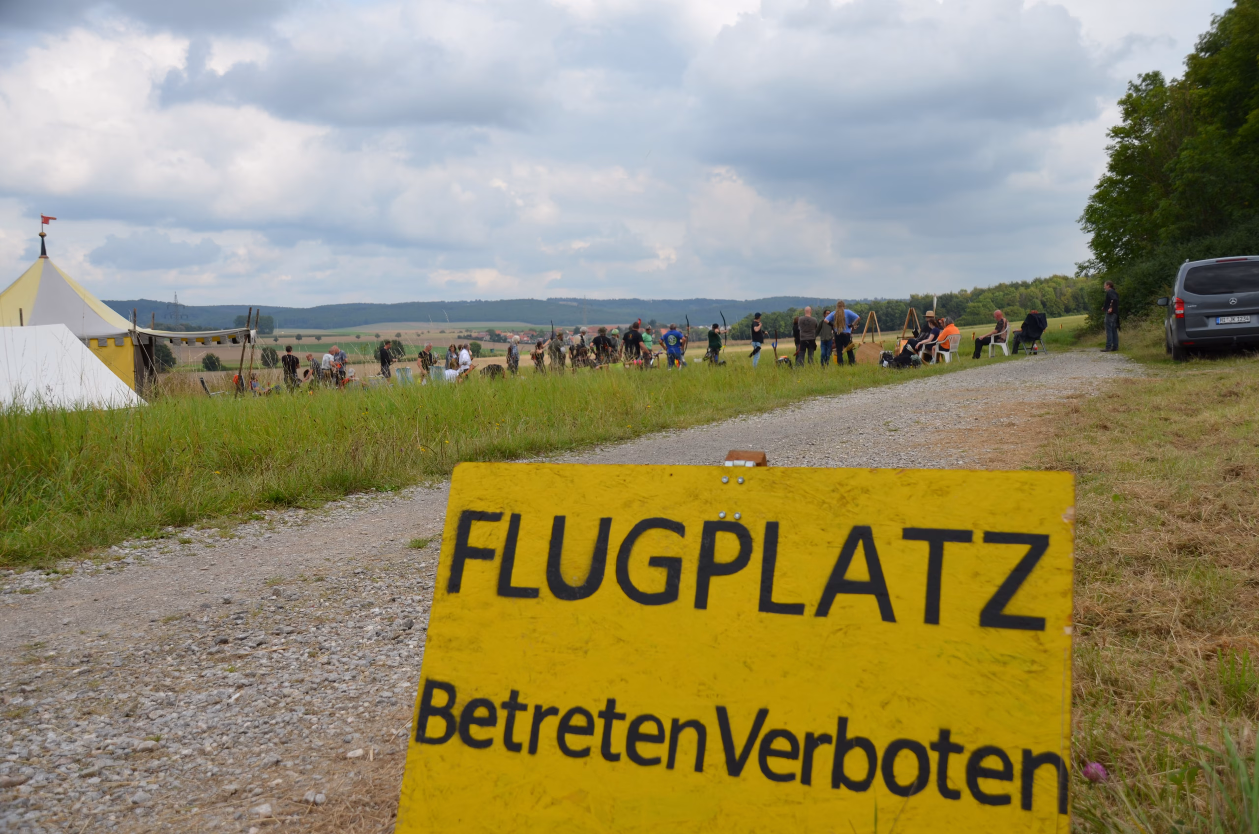 Gelbes Warnschild 'Flugplatz Betreten Verboten' vor dem Siebenberge Clout Bogenwettbewerb 2021 von Pfeilflugkunst mit Bogenschützen und Zelten in der Landschaft.