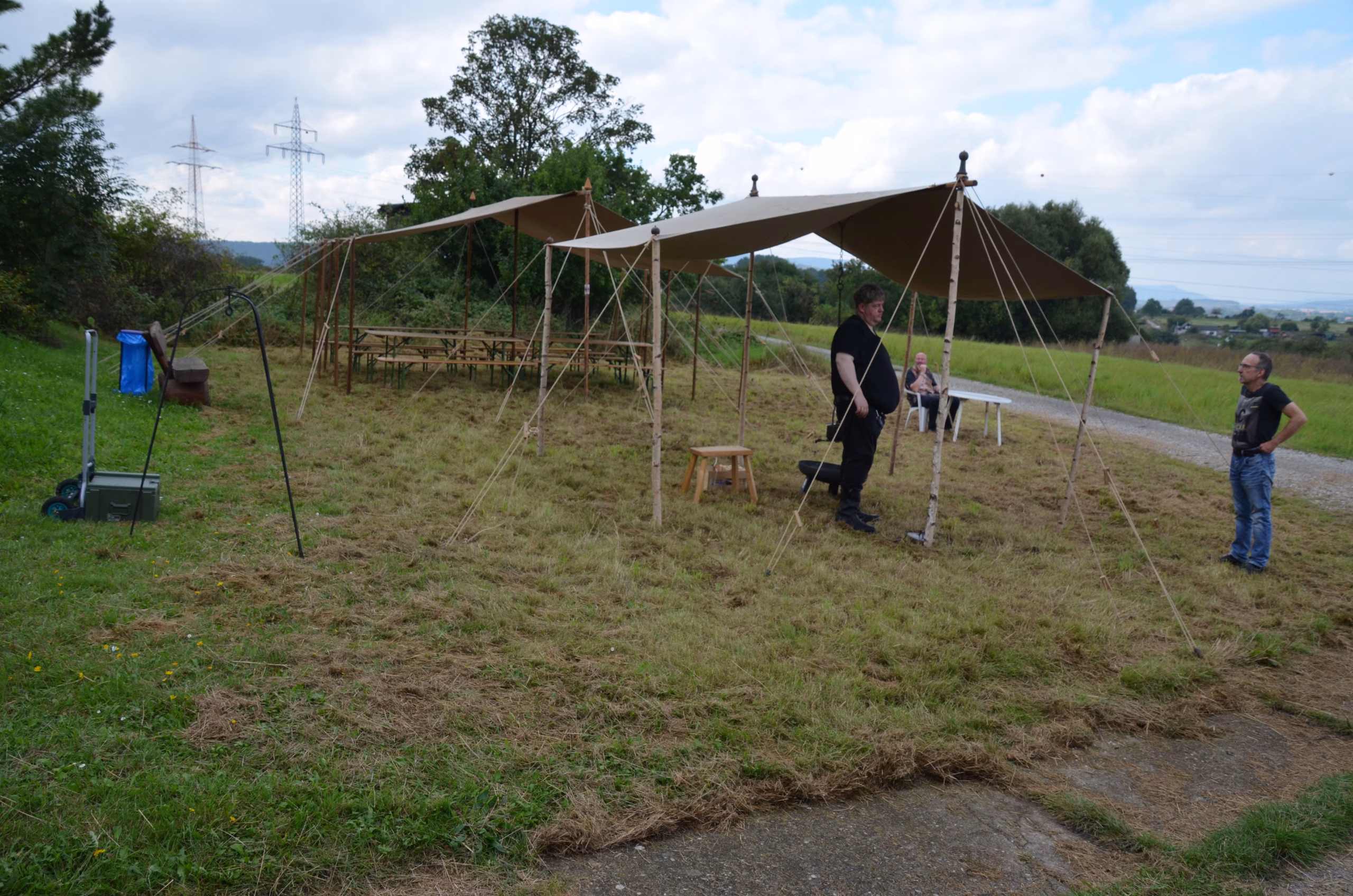 Zwei Männer unter einem rustikalen Zelt bei einem Clout-Bogenschieß-Wettbewerb von Pfeilflugkunst in Siebenberge, Leinebergland, mit weiteren Zelten und Tischen im Hintergrund.