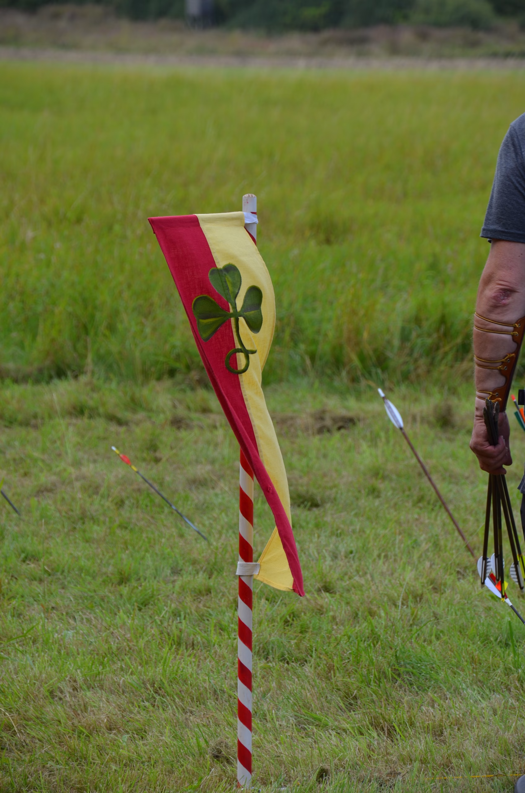 Rot-gelbe Ziel-Flagge mit Kleeblatt beim Siebenberge Clout Bogenwettbewerb, daneben ein Bogenschütze mit Pfeilen.