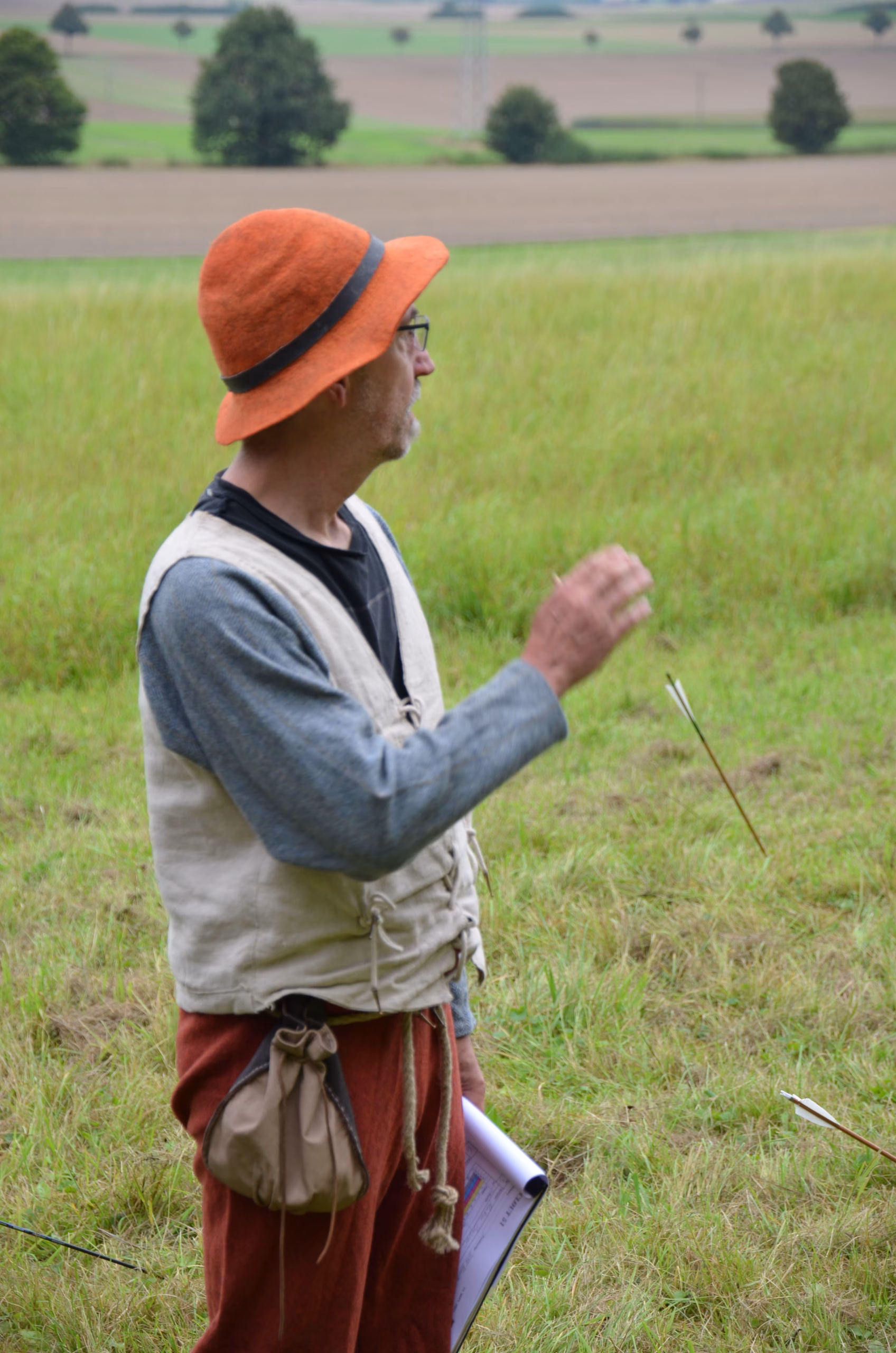 Mann mit orangefarbenem Hut und traditioneller Kleidung beim Siebenberge Clout Bogenschießen, Pfeile im Gras.