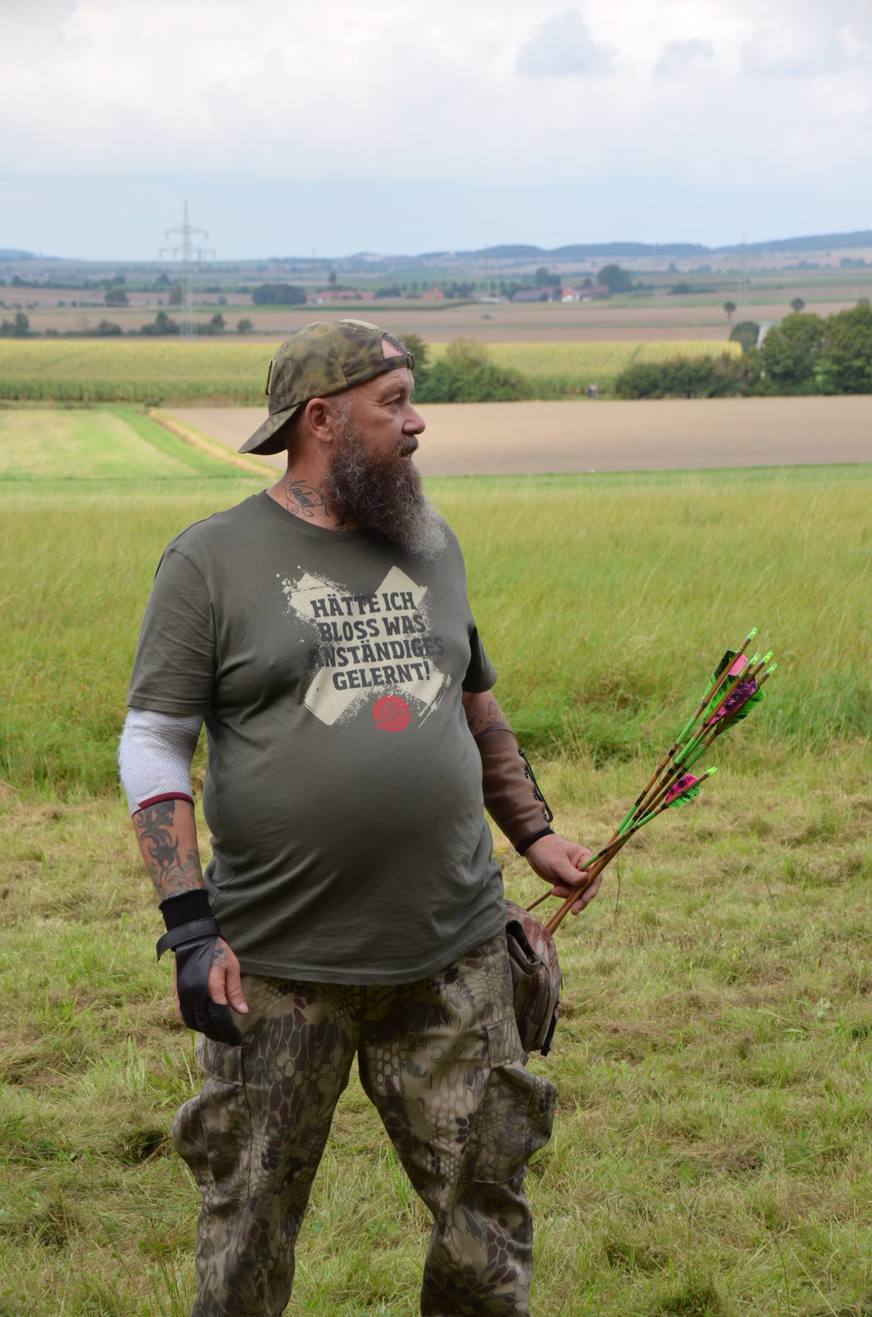 Bogenschütze mit Bart und Tattoos, der Pfeile hält, beim Siebenberge Clout Bogenwettbewerb 2021 von Pfeilflugkunst in einer weiten Feldlandschaft.
