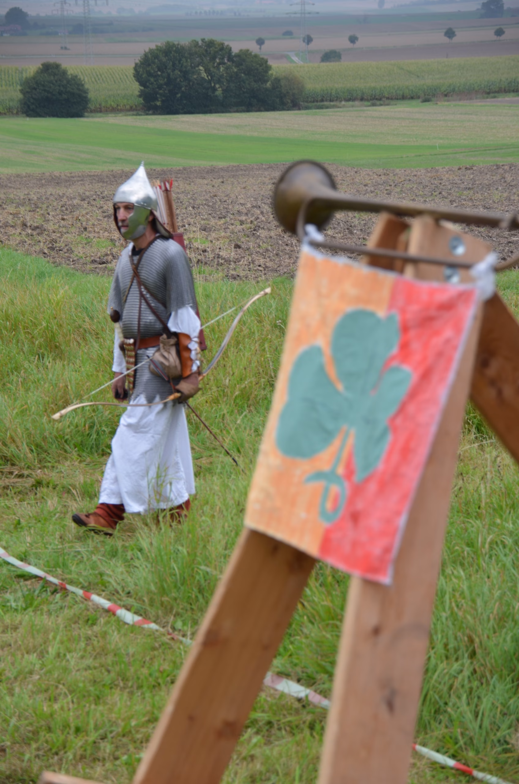 Bogenschütze in historischer Rüstung beim Siebenberge Clout Bogenwettbewerb von Pfeilflugkunst, mit Clout-Flagge im Vordergrund.