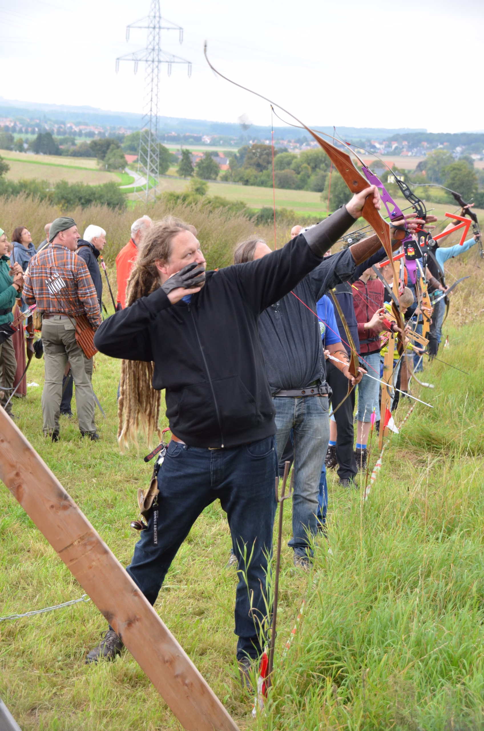 Ein Bogenschütze mit Dreadlocks spannt seinen Recurvebogen beim Clout-Bogenschießen in Siebenberge, veranstaltet von Pfeilflugkunst.