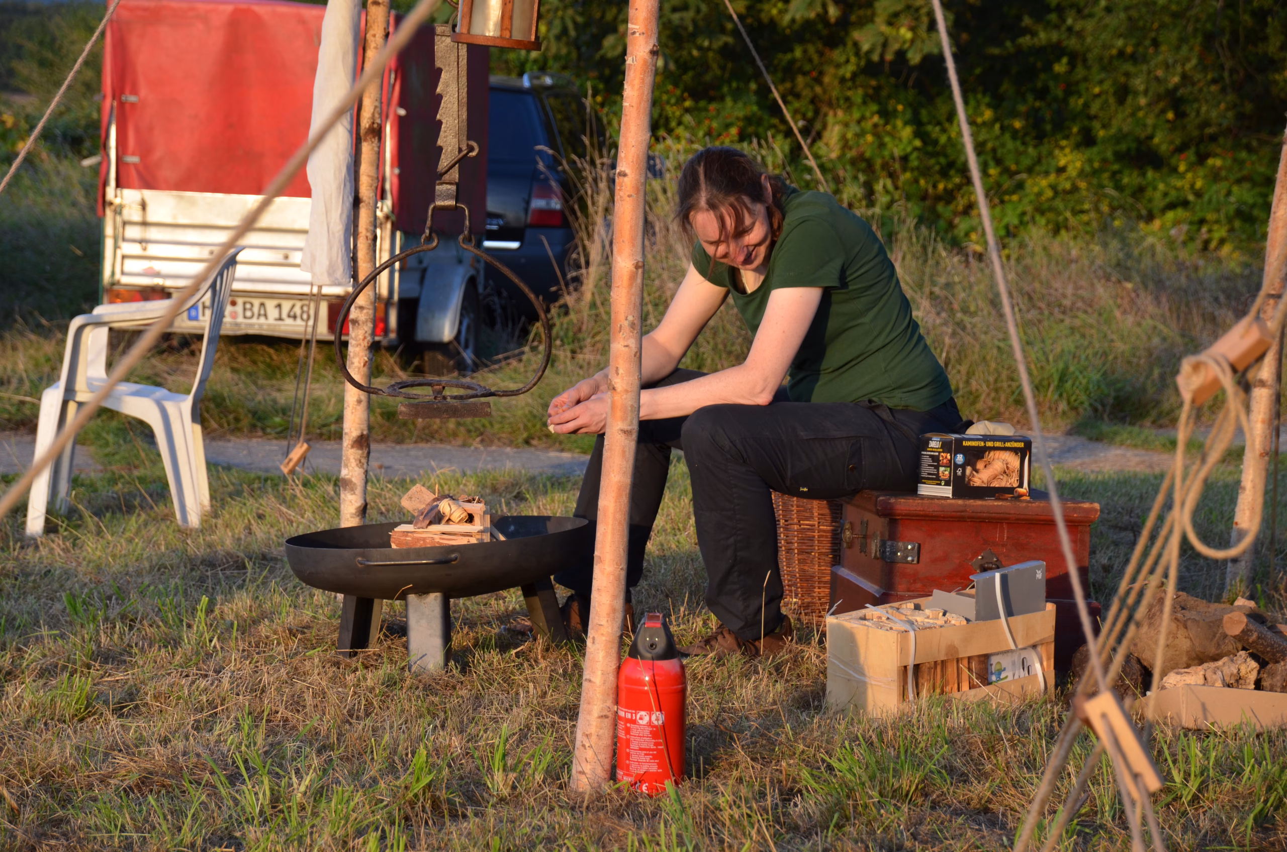 Eine lächelnde Bogenschützin sitzt entspannt neben einer Feuerschale, bereit für ein Lagerfeuer, beim Siebenberge Clout Bogenwettbewerb von Pfeilflugkunst.