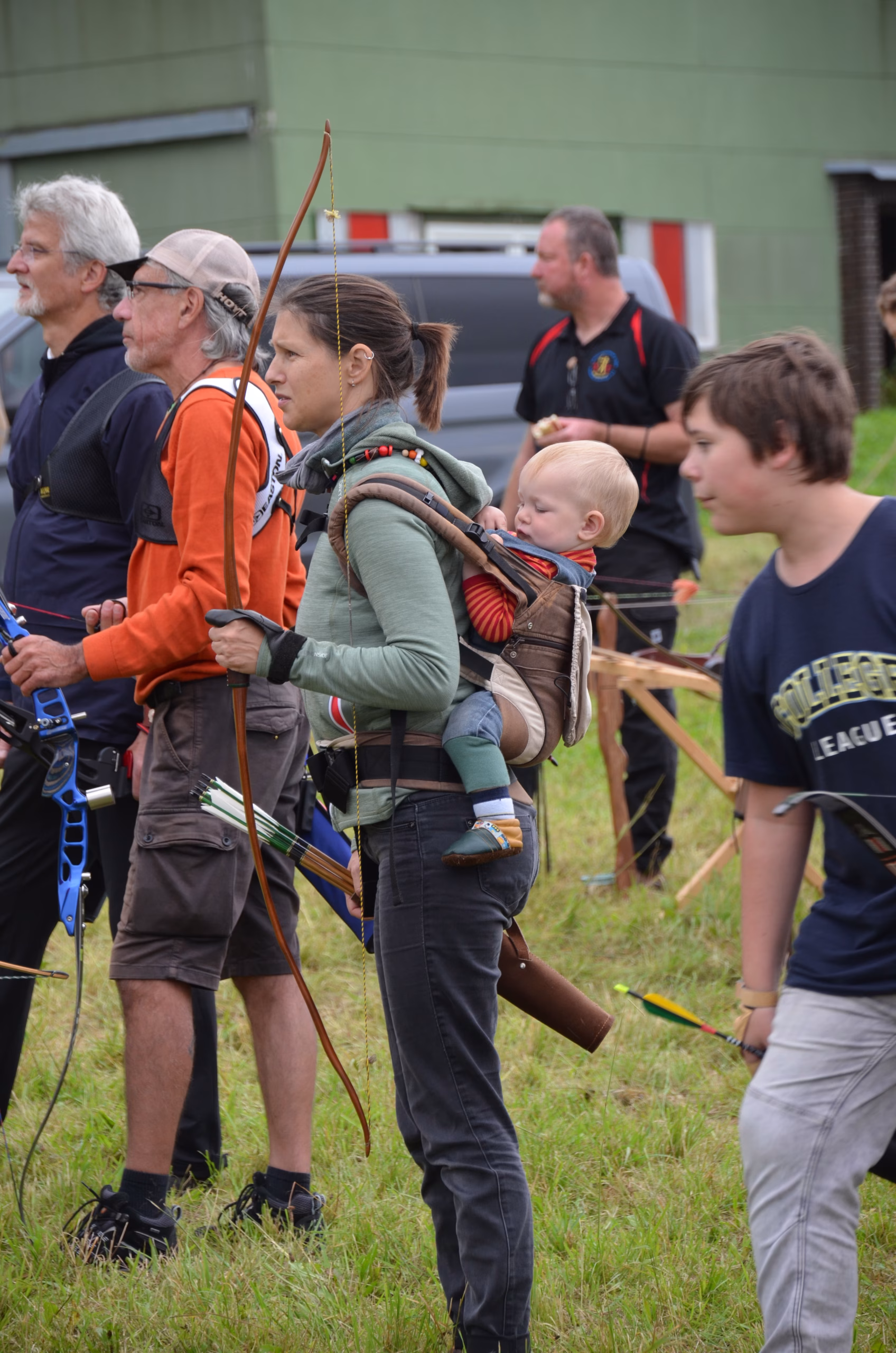 Eine Bogenschützin mit traditionellem Bogen und Baby in einer Trage beim Clout-Bogenschießen in Siebenberge, veranstaltet von Pfeilflugkunst.