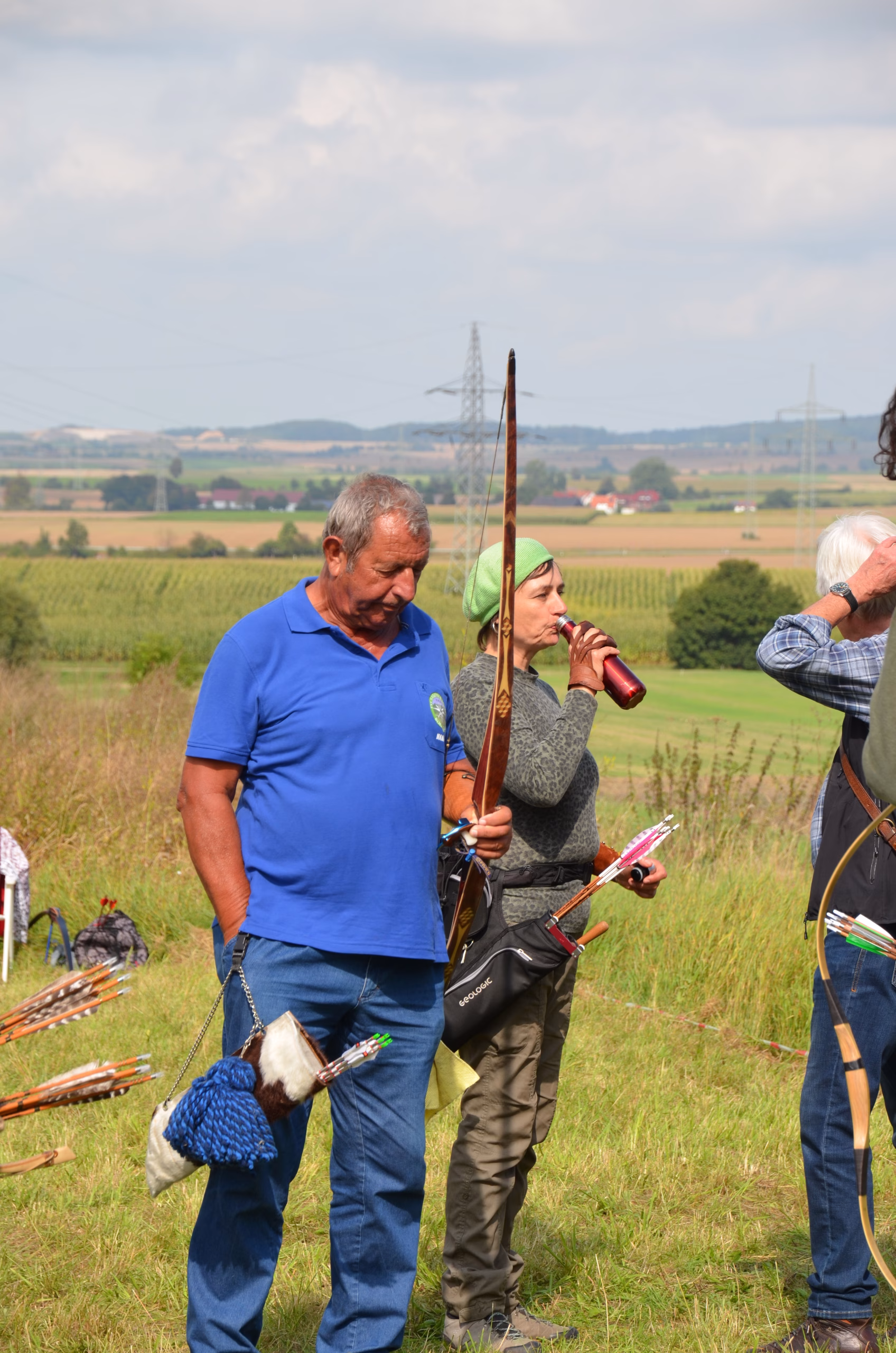 Zwei Bogenschützen mit traditionellen Bögen und Köchern machen eine Pause beim Siebenberge Clout Bogenwettbewerb 2021 von Pfeilflugkunst in einer weiten Feldlandschaft.