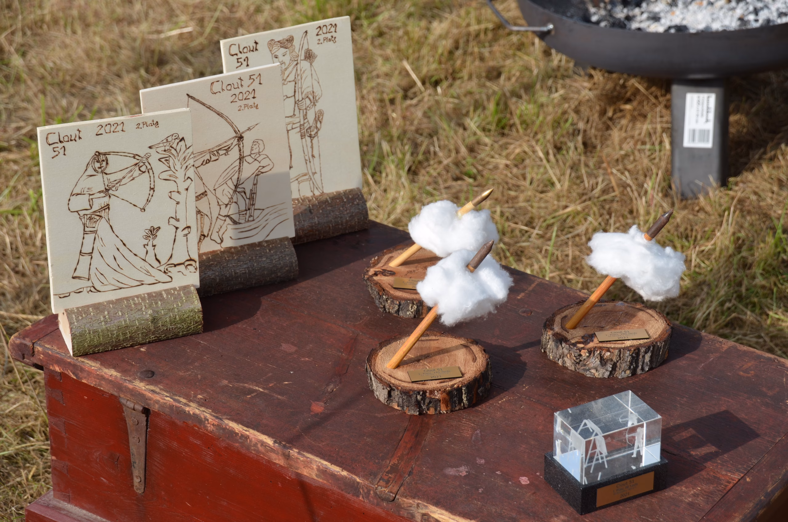 Handgefertigte Preise und Trophäen des Siebenberge Clout Bogenwettbewerbs 2021 von Pfeilflugkunst, darunter Holztafeln mit Brandmalerei und kreative Pfeil-Wolken-Trophäen.