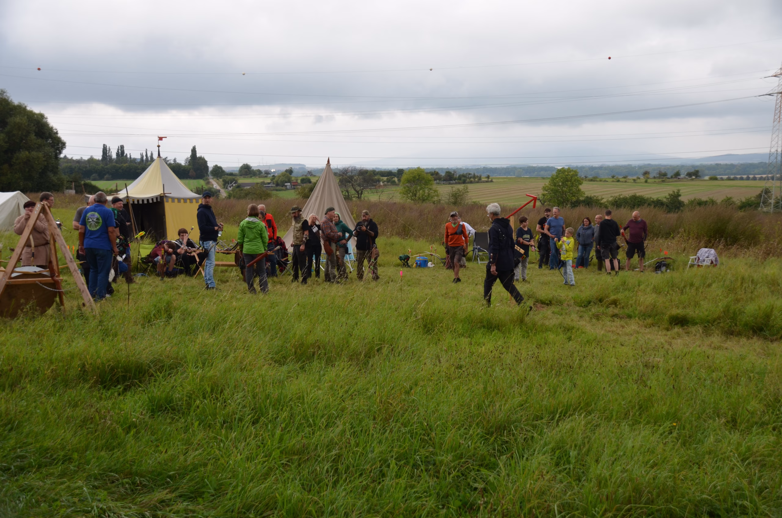 Bogenschützen und Zuschauer beim Siebenberge Clout Bogenwettbewerb 2021 von Pfeilflugkunst in einem grünen Feld mit Zelten im Hintergrund.