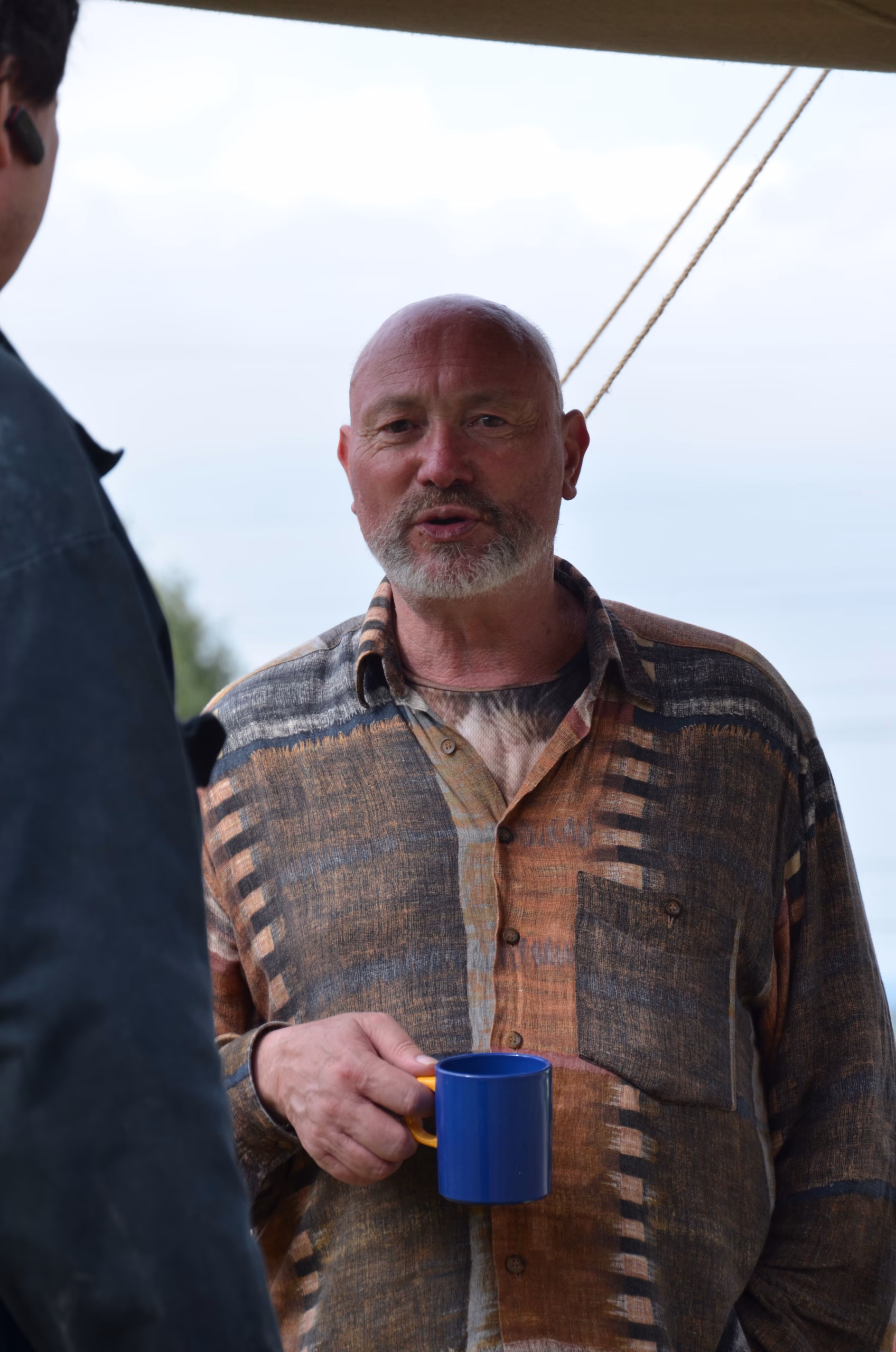 Mann mit Bart und gemustertem Hemd hält blaue Tasse, im Gespräch beim Siebenberge Clout Bogenwettbewerb von Pfeilflugkunst.